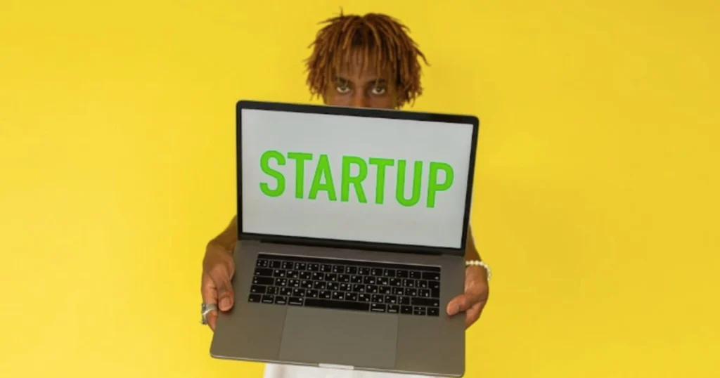 Ragazzo in primo piano che sorregge un computer con la scritta Startup in un ambiente di coworking